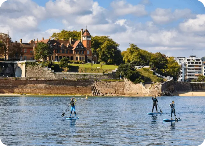 Personas practicando paddle surf en la bahía de la Concha, con el Palacio Miramar al fondo.